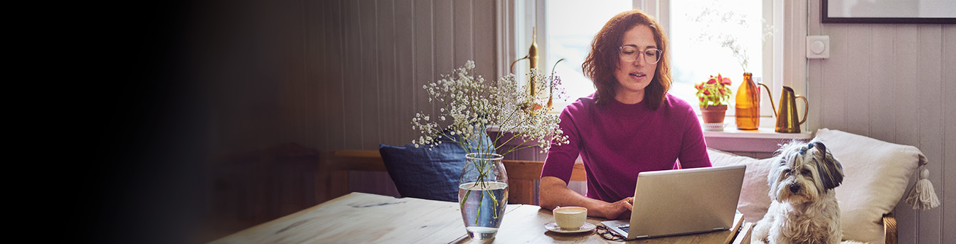 Kvinne med laptop og kaffekopp sitter ved spisebordet på hytta sammen med hunden sin. Blomstervase på bordet.