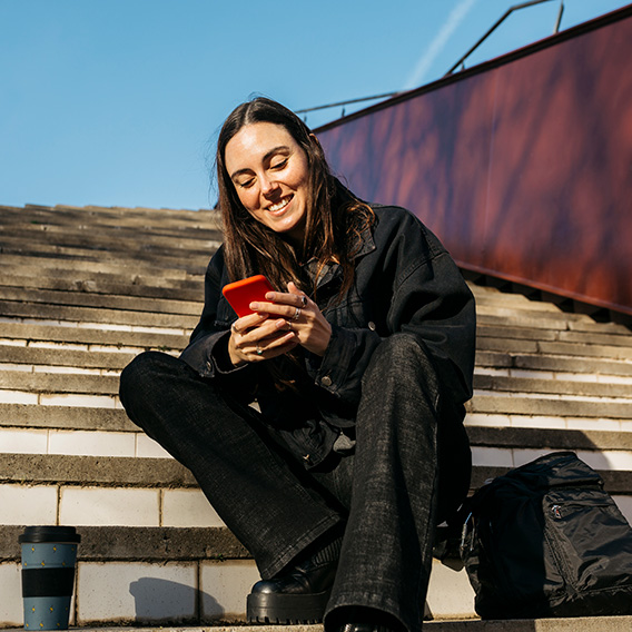 Ung, smilende kvinne med brunt hår sitter i trappa i parken og tekster med sin røde smarttelefon.